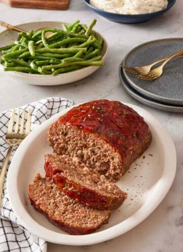 Sliced meatloaf on a serving platter. Green beans, plates, flatware, and mashed potatoes in the background.