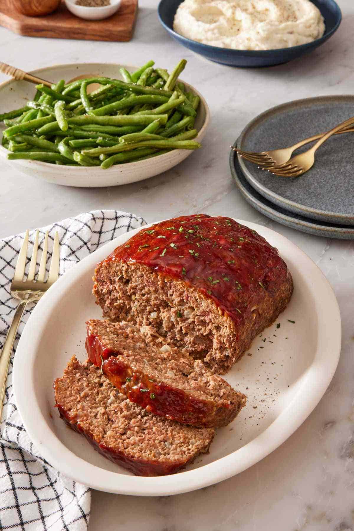 Sliced meatloaf on a serving platter. Green beans, plates, flatware, and mashed potatoes in the background.