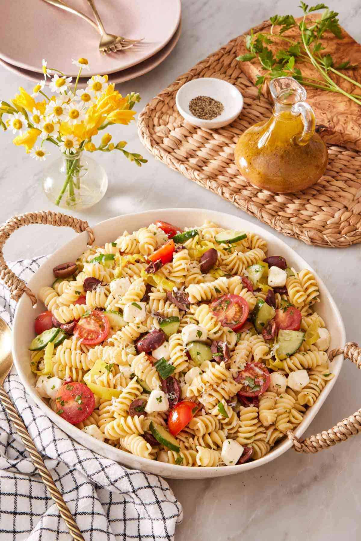 A deep dish of pasta-based salad with a small vase of flowers alongside. A small jar of salad dressing is next to it.