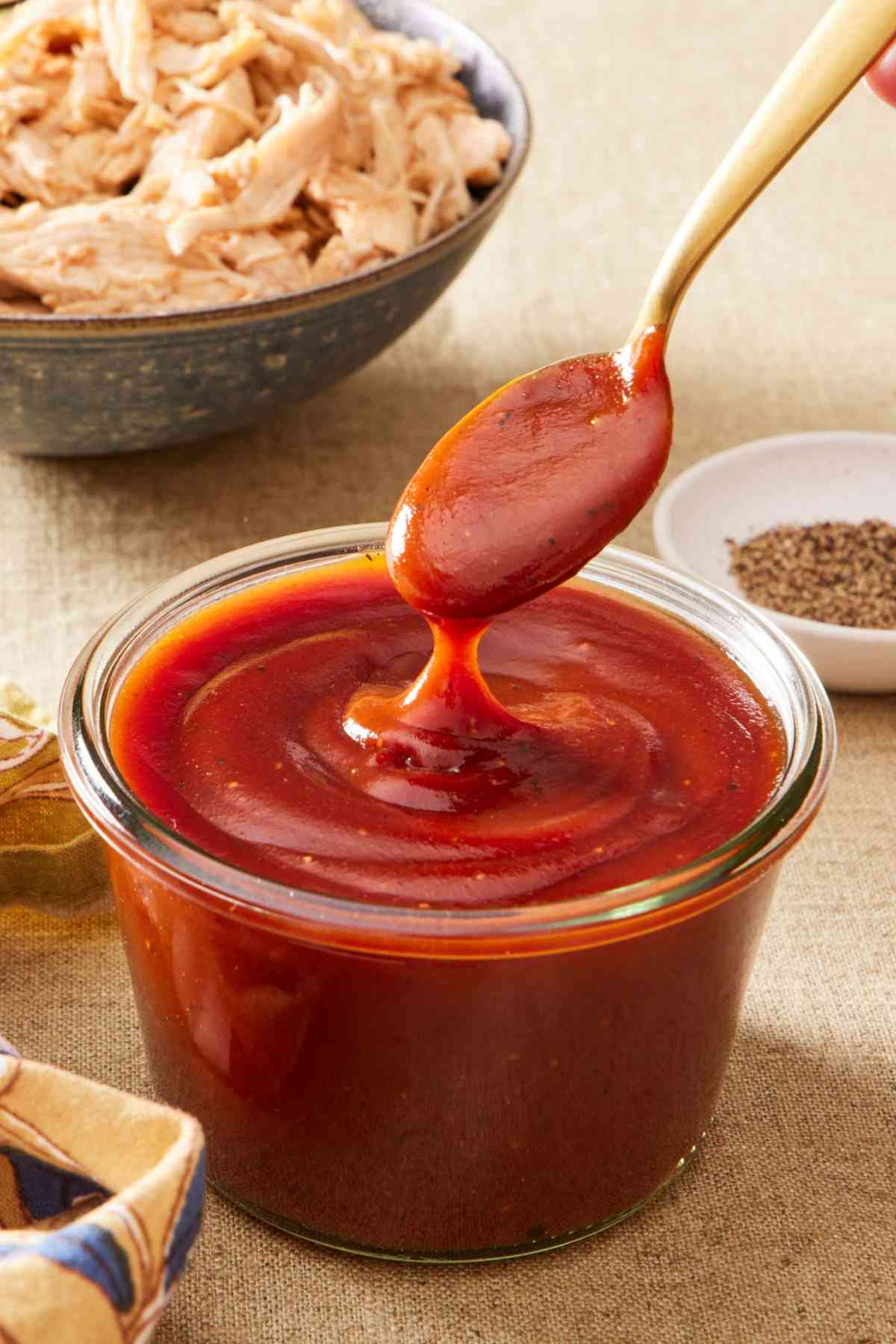 A spoon dipped into a small jar of homemade BBQ sauce. Shredded meat and black pepper in the background.