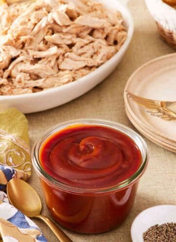 Homemade barbecue sauce recipe in a small glass container with a spoon next to it. Shredded meat, black pepper, and plates around it.