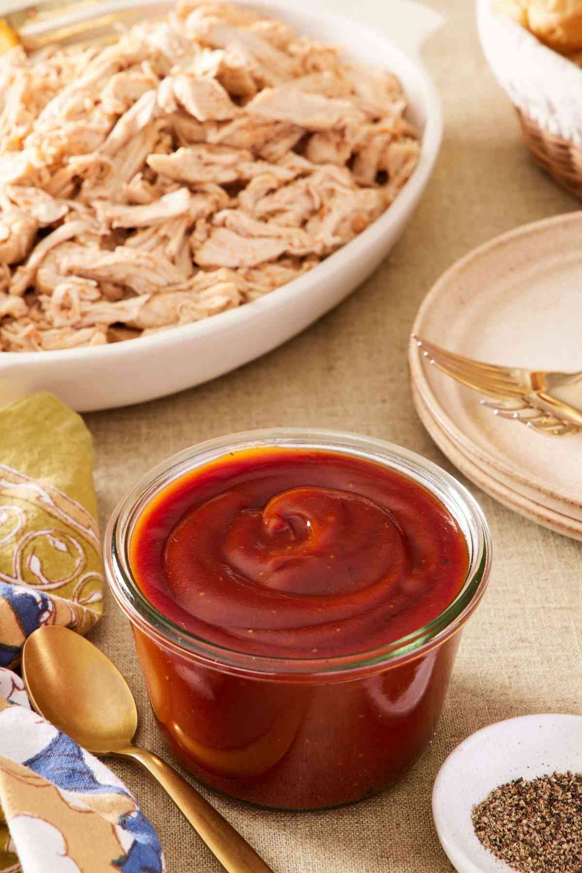 Homemade barbecue sauce recipe in a small glass container with a spoon next to it. Shredded meat, black pepper, and plates around it.