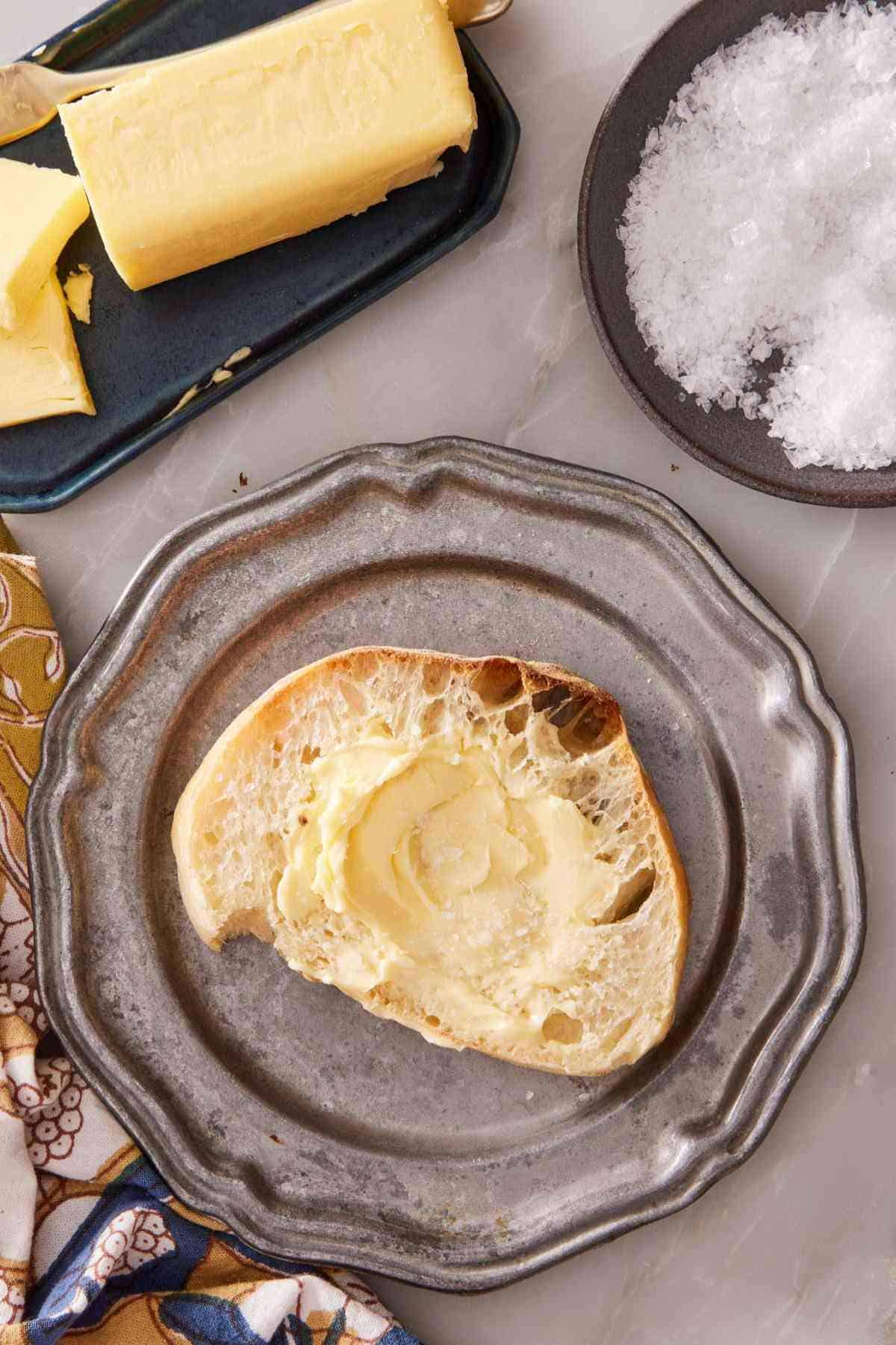 A buttered slice of homemade ciabatta bread on a plate with a stick of butter and a small dish of salt on the side.