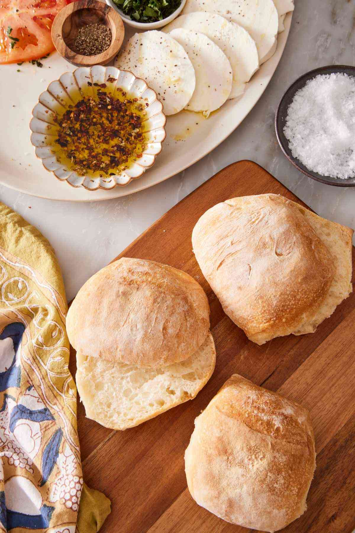 Ciabatta rolls on a wooden board with a small dish of salt alongside, and a platter of herbed olive oil and sliced fresh mozzarella in the background.