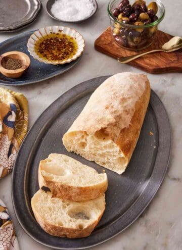 A loaf of ciabatta bread with two slices cut out, with olives and olive oil in the background.