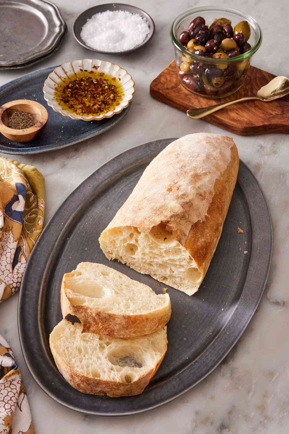 A loaf of ciabatta bread with two slices cut out, with olives and olive oil in the background.