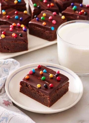 Cosmic brownie on a white plate next to a side of milk, with a platter of them in the background.