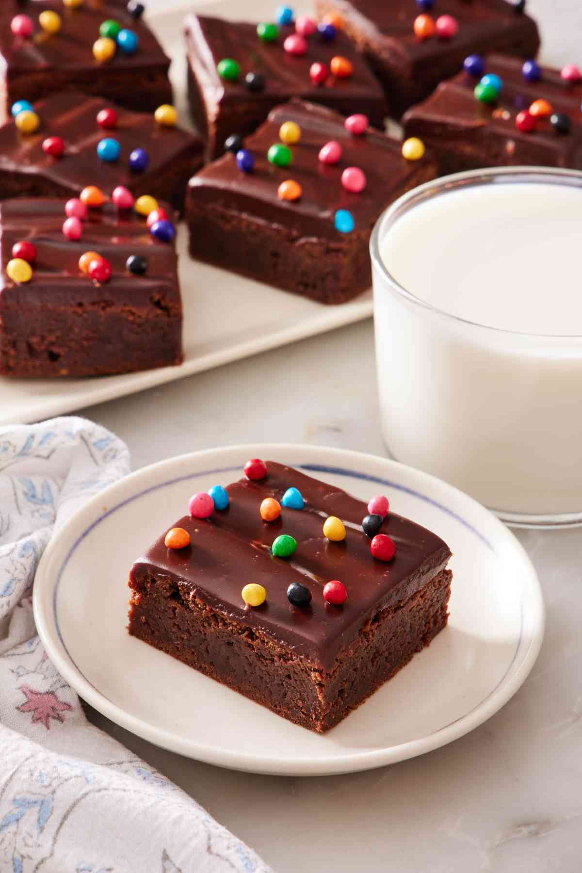 Cosmic brownie on a white plate next to a side of milk, with a platter of them in the background.