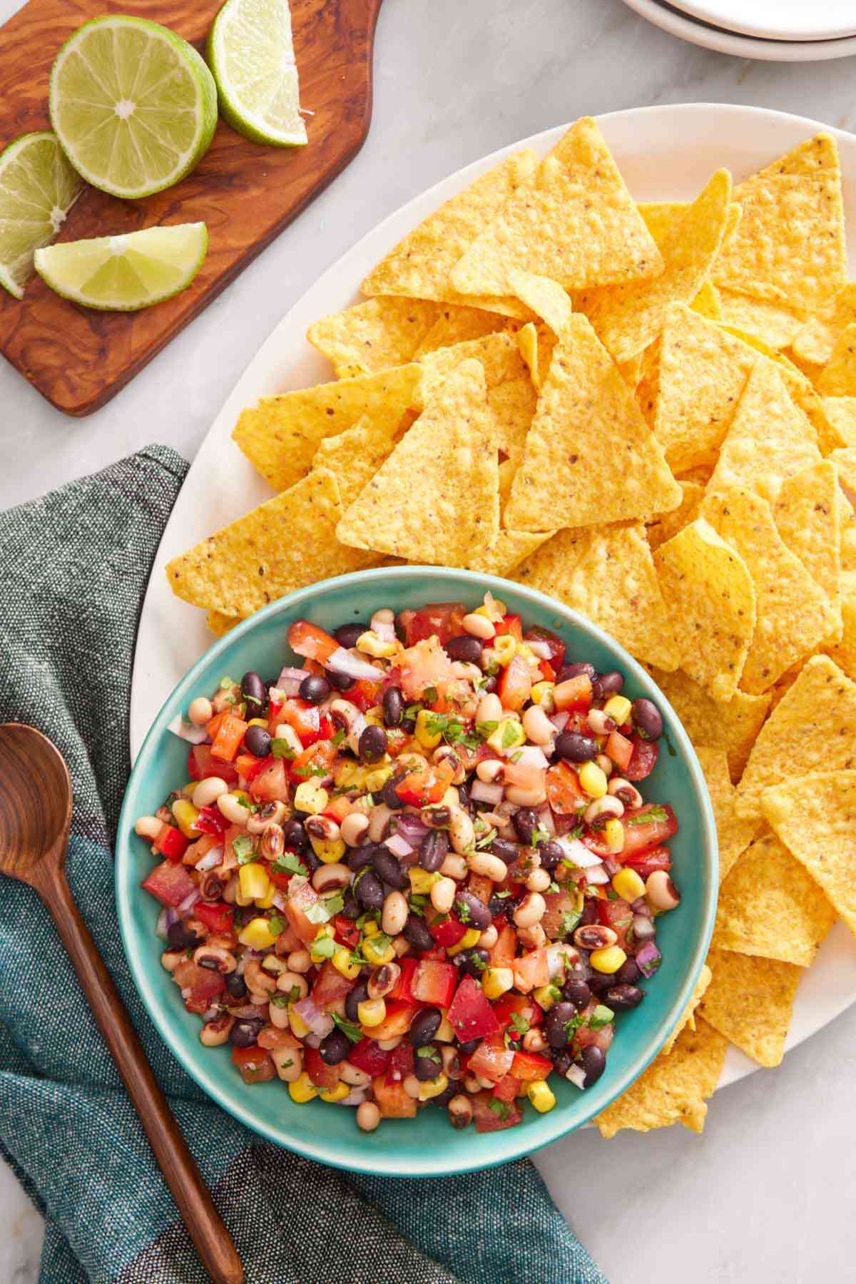 Overhead shot of a bowl of cowboy caviar set on a platter with chips. Lime on a wooden board off to the side,