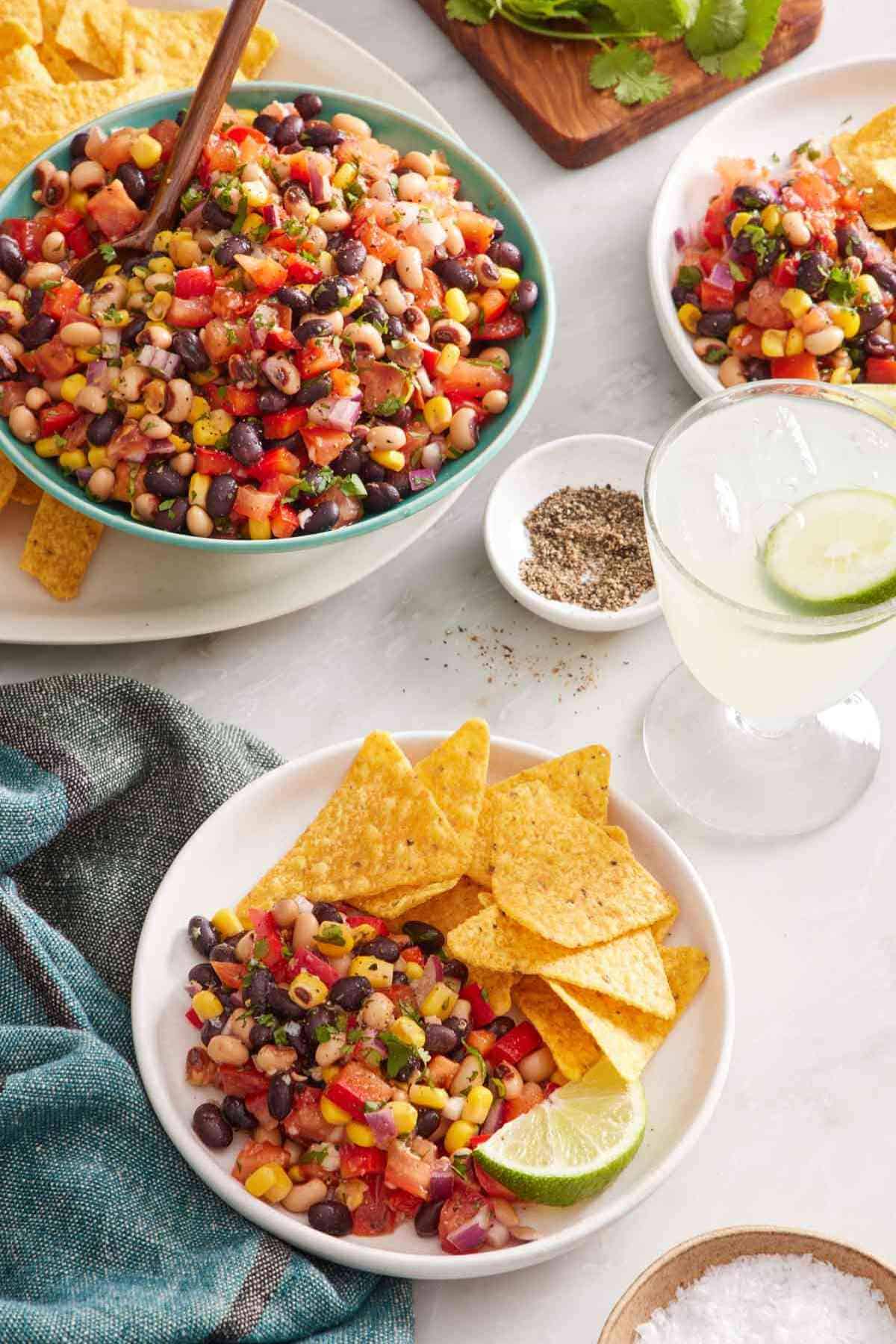 A serving of cowboy caviar with chips and lime in a bowl. A drink is on the side, with the rest of the dish in the background.