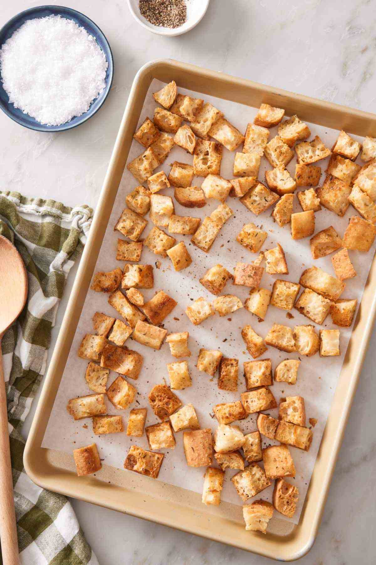 Overhead shot of homemade croutons on a lined baking sheet with a small dish of salt and another of pepper alongside.