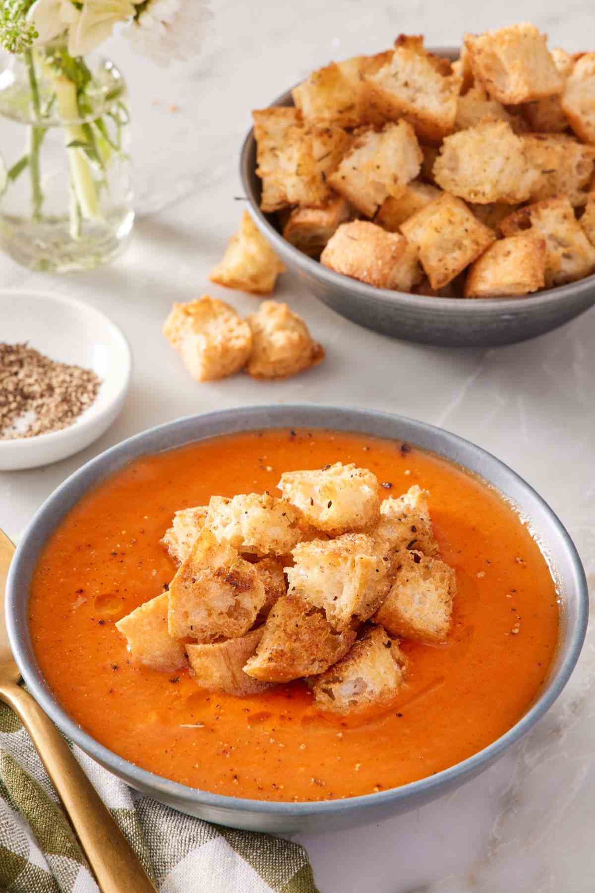 A bowl of soup topped with croutons, with another bowl of croutons only in the background.