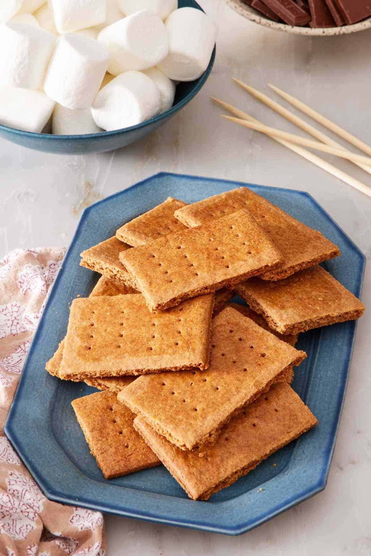 Graham crackers on a blue platter with skewers, marshmallows, and chocolate in the background.