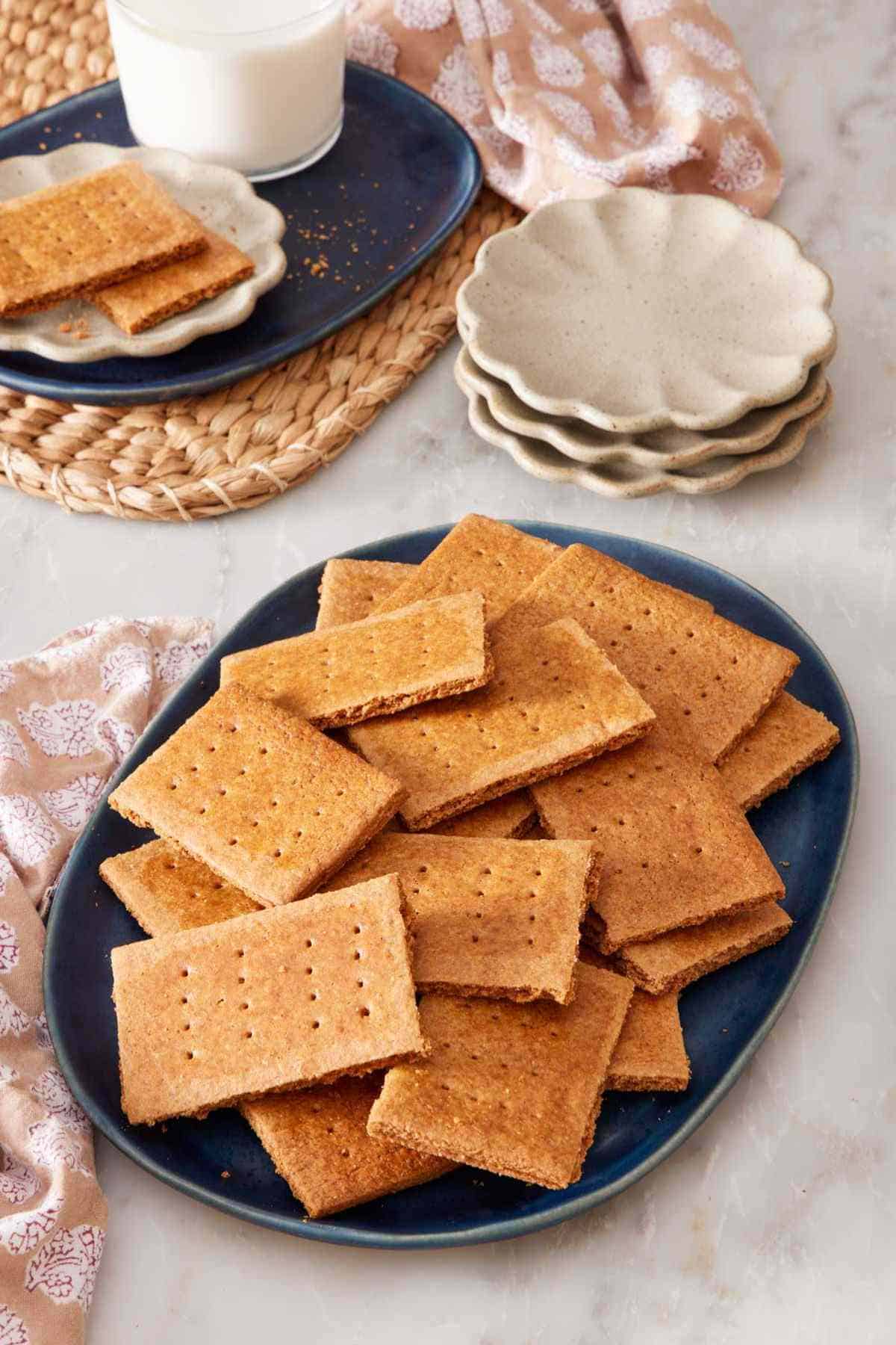 Graham crackers on a blue platter with small serving plates, more crackers, and a glass of milk in the background.