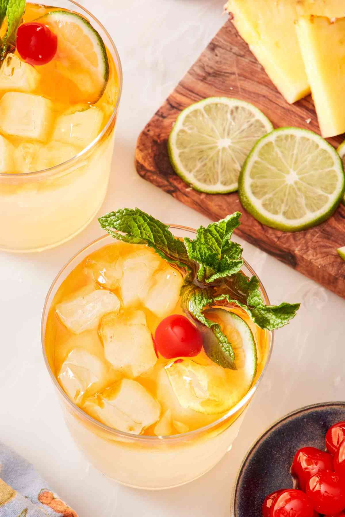 Overhead shot of a mai tai cocktail and a second cocktail slightly cut off. A small dish of cherries and a board with lime wheels and pineapple wedges around the glass.