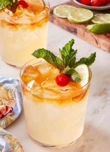 Easy mai tai recipe garnished with lime, mint, and cherry. Another cocktail and a wooden board with cocktail garnishes in the background.