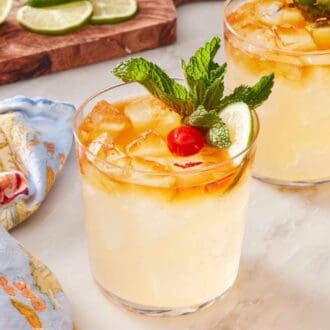 Two mai tais on a marble countertop with a table linen on the side, as well as a small dish of cherries. Wooden board with lime wheels and wedges in the background.