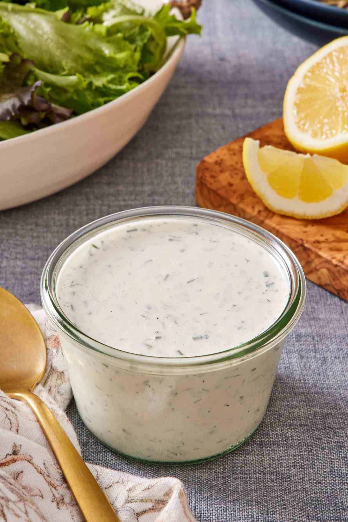 Ranch dressing in a small glass jar with sliced lemons and greens in the background.