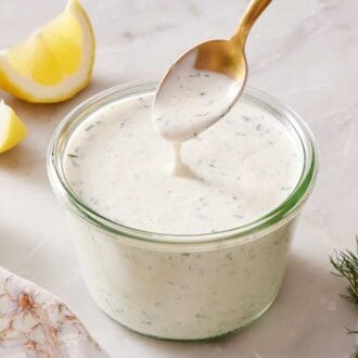 A spoon dipped into homemade ranch in a glass jar, with sliced lemon and fresh dill on the side.