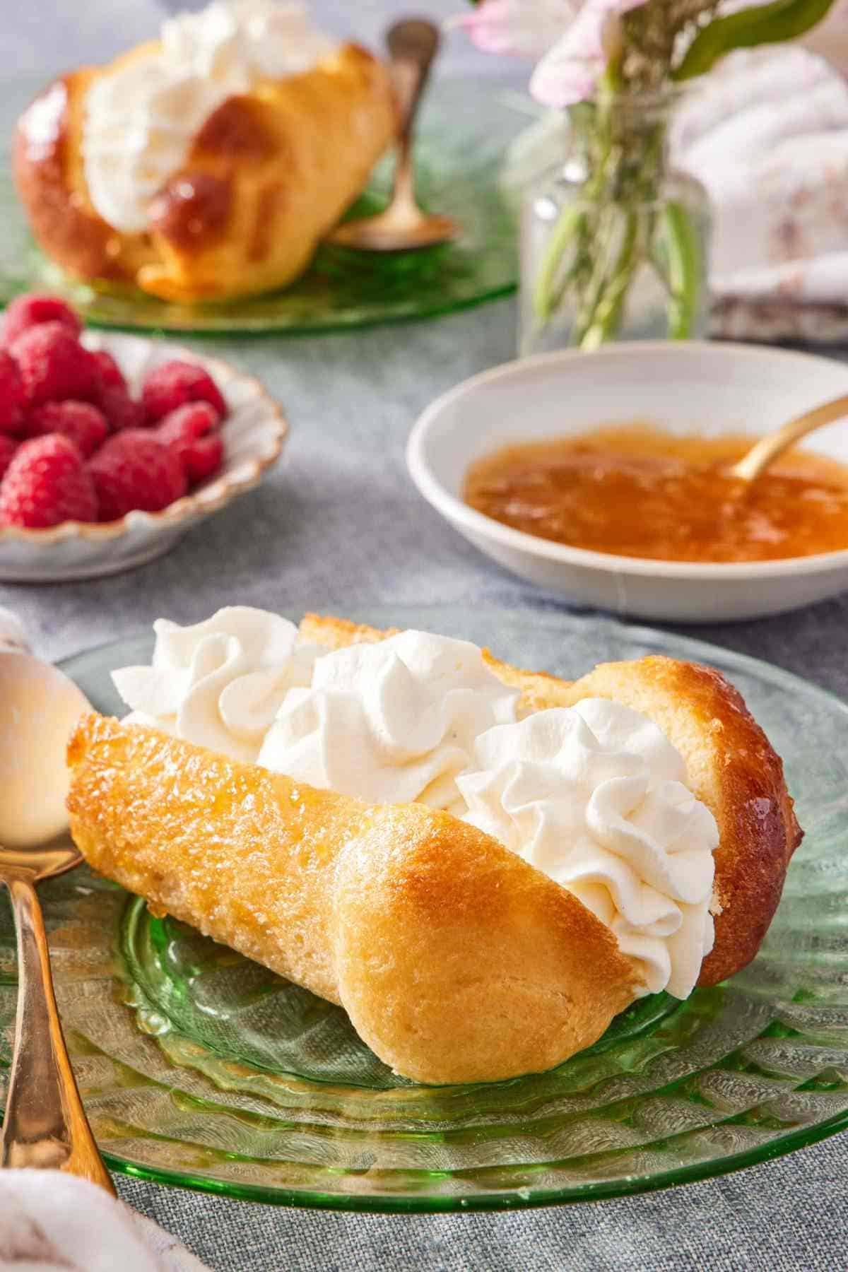 A rum baba halved and filled with whipped cream. Syrup, berries, and another filled rum baba in the background.