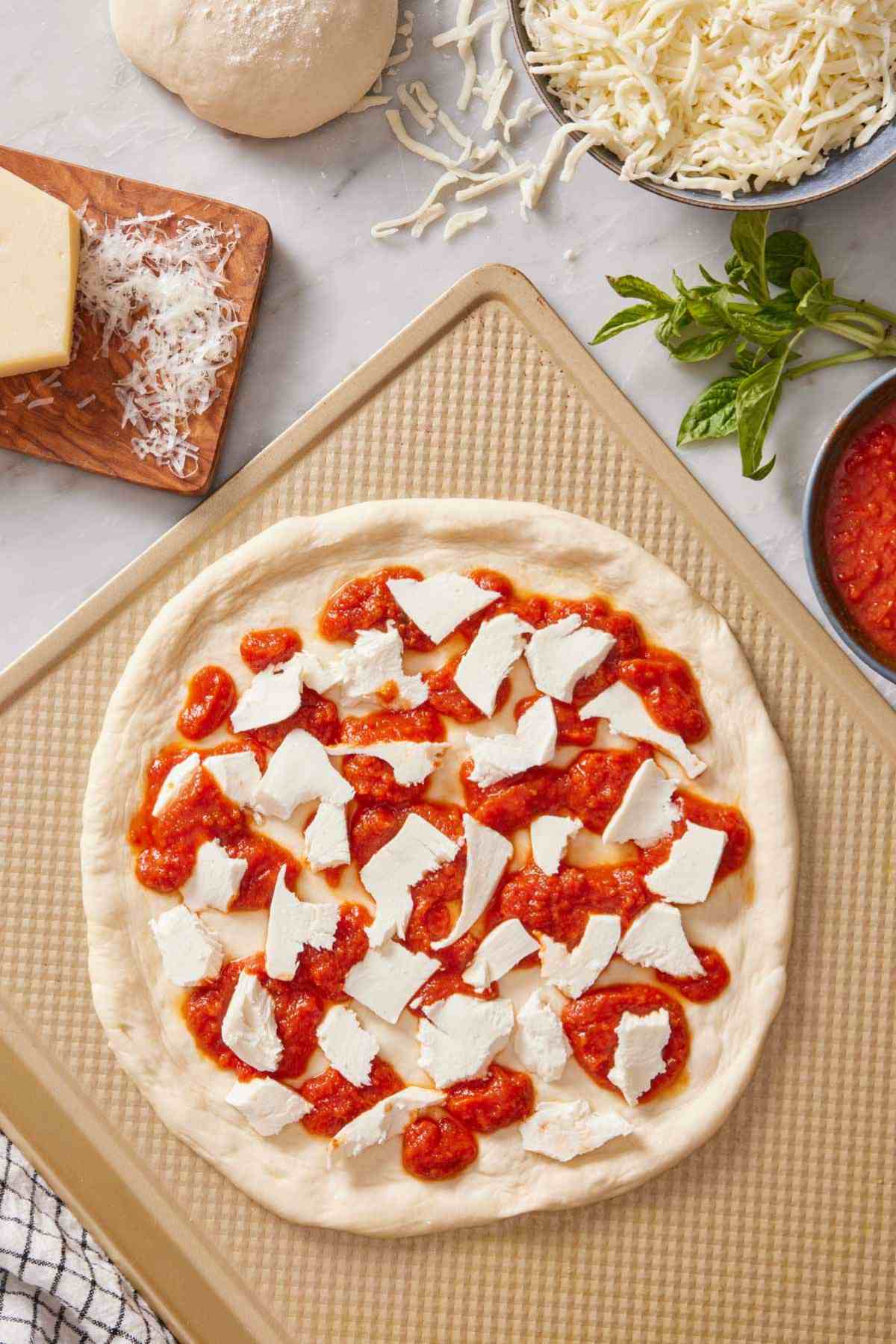 Overhead shot of sourdough pizza crust on a baking sheet with pizza sauce and torn cheese on top. More pizza toppings around it.