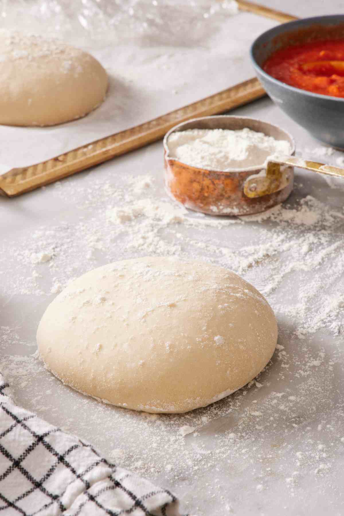 Sourdough pizza dough on a lightly floured surface with a measuring cup of flour, more dough, and pizza sauce in the background.