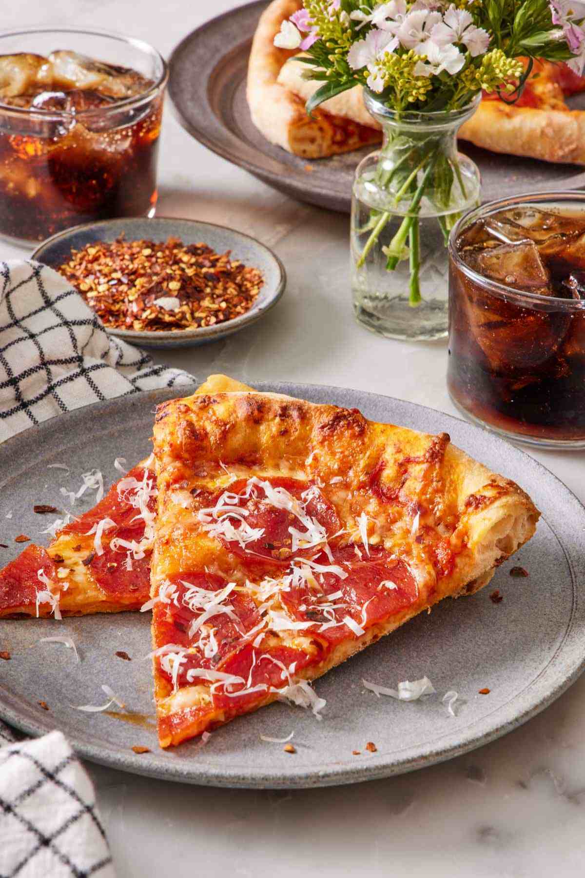 A slice of pizza with pepperoni and shredded cheese on top. Two drinks, a small dish of chili flakes, a vase of flowers, and more pizza in the background.