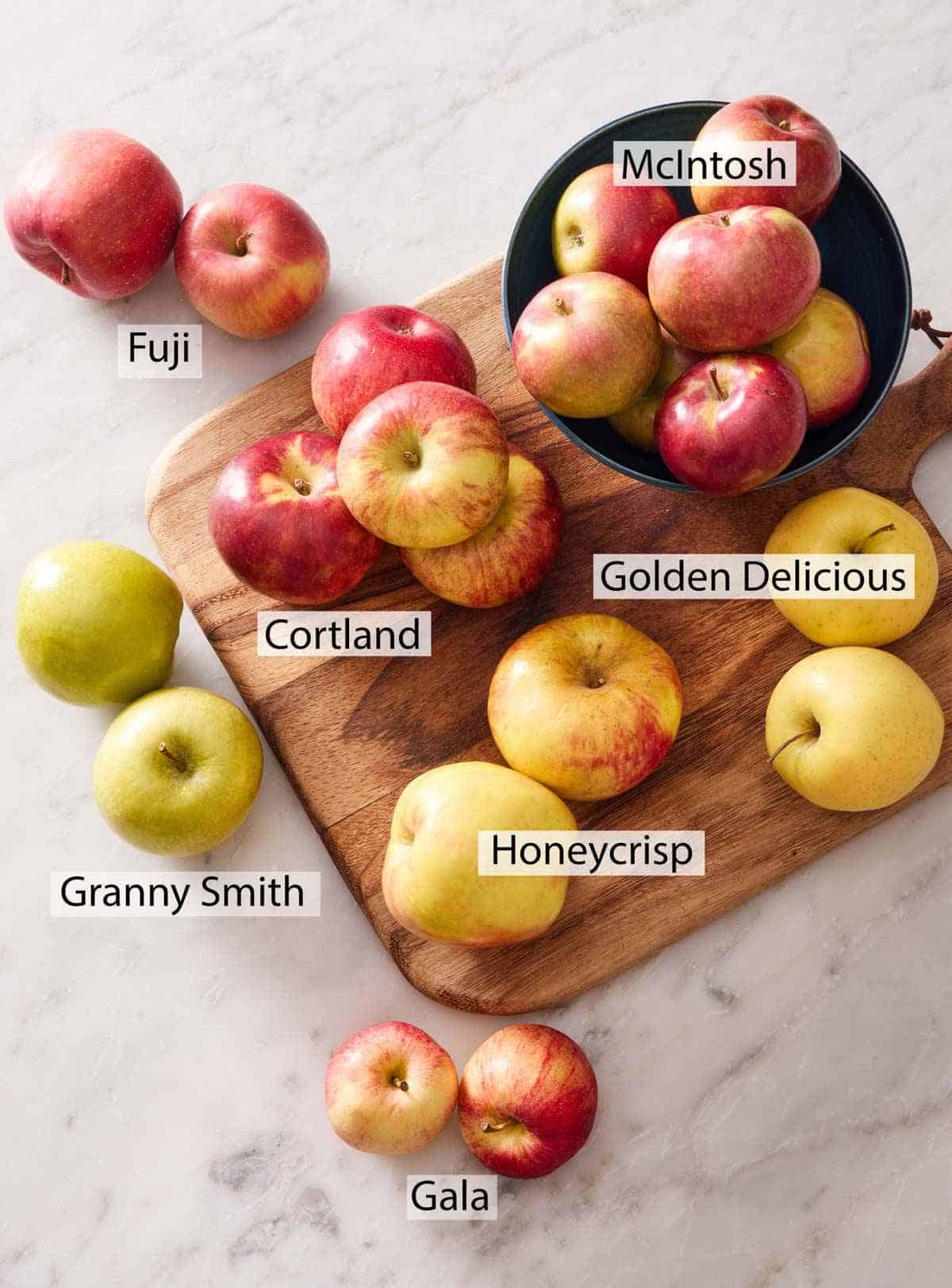 Various types of apples scattered and piled on a wooden board and in a bowl.
