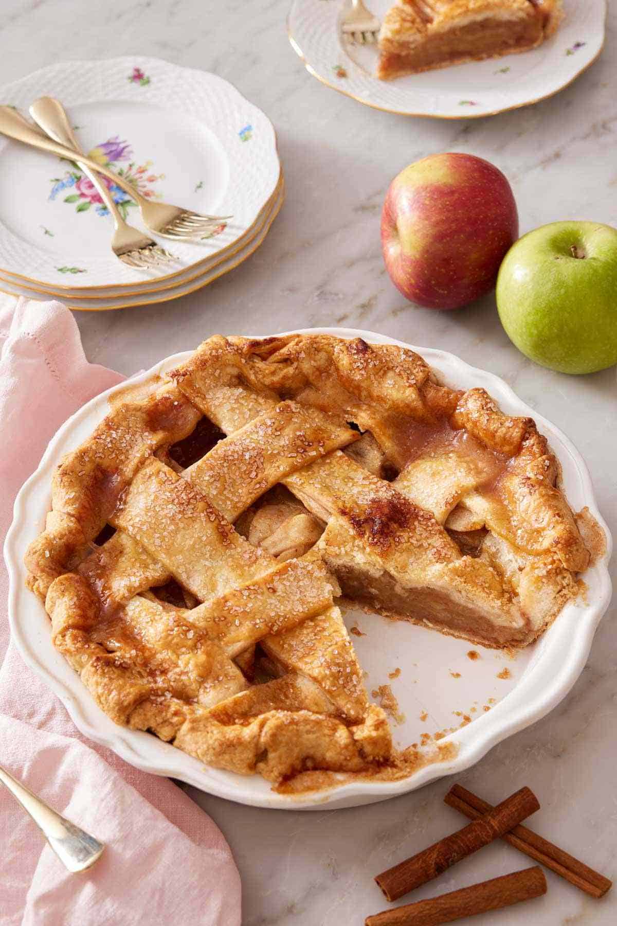 Homemade apple pie recipe with a lattice top with a slice cut out. Some apples, cinnamon sticks, and a few small plates around it.
