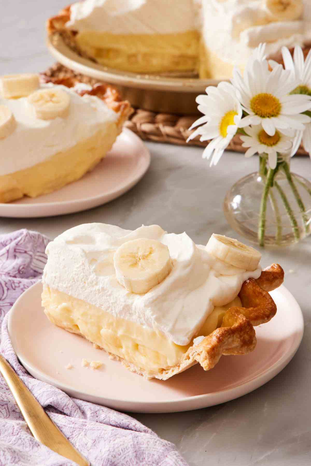 A slice of banana cream pie on a plate with more pie and a small vase of flowers in the background.