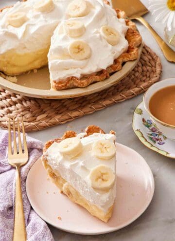 A slice of banana cream pie next to a cup of tea, with the rest of the pie in the background.