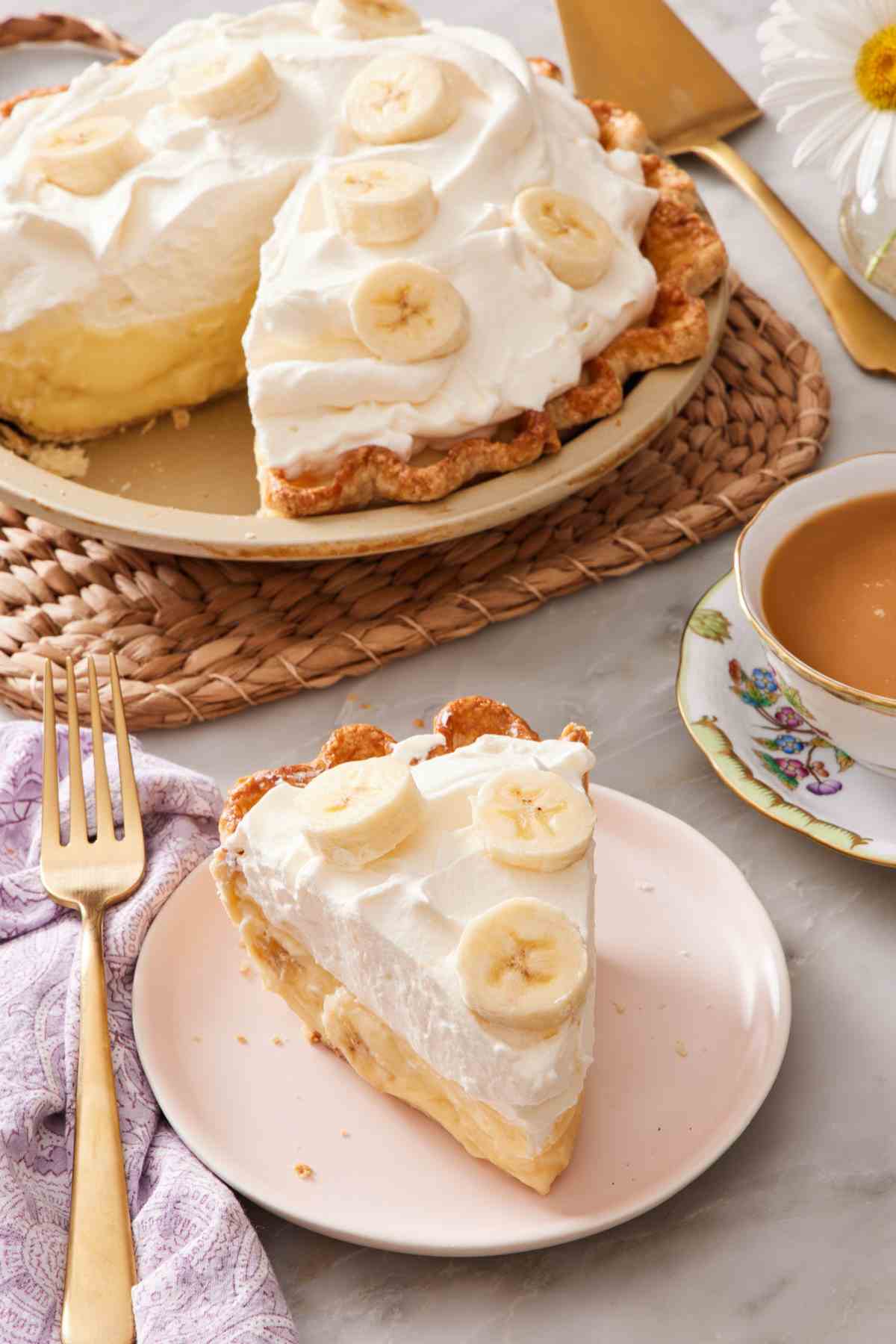 A slice of banana cream pie next to a cup of tea, with the rest of the pie in the background.