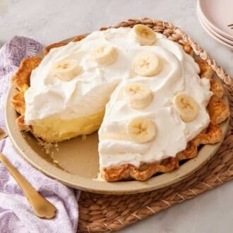 Banana cream pie with a slice cut out of it. A cake server rests on a purple table linen alongside.