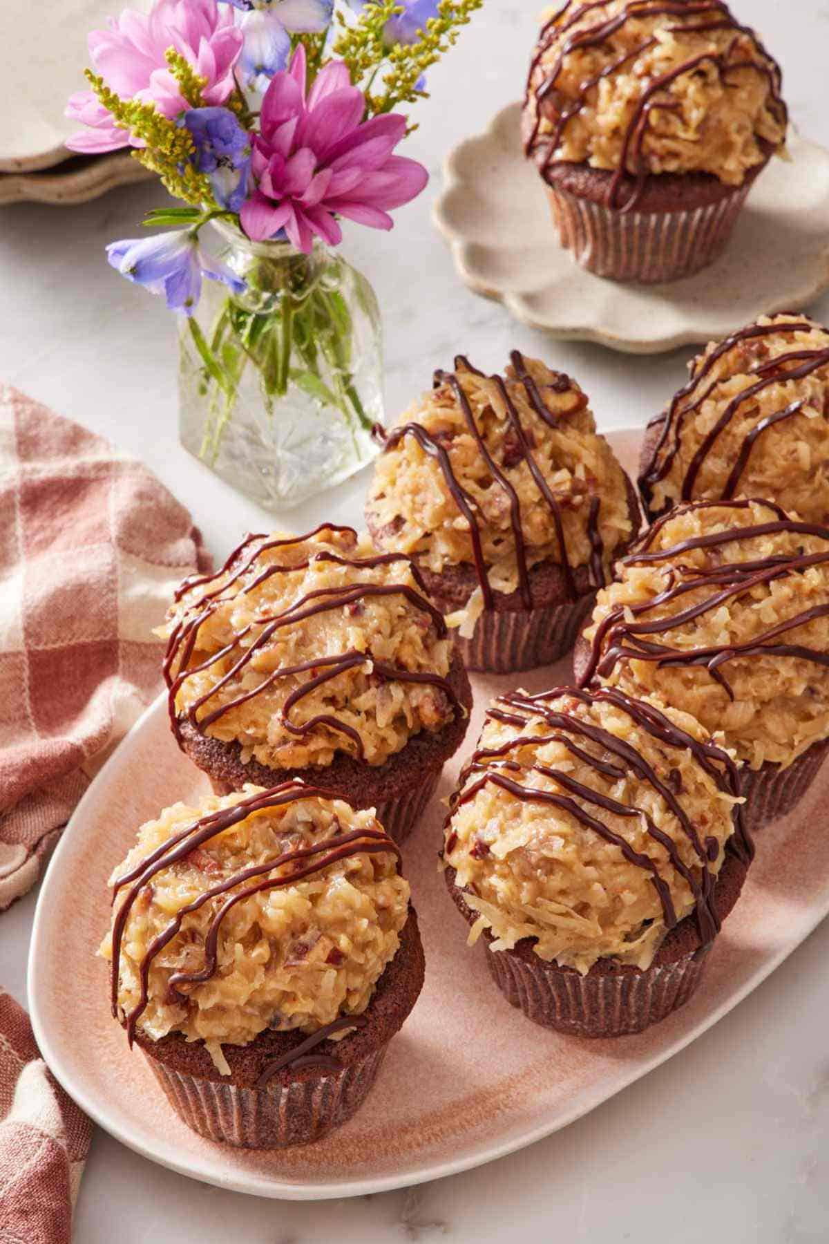 A platter of German chocolate cupcakes with a small vase of flowers alongside. A single cupcake sits on a small serving plate in the background.