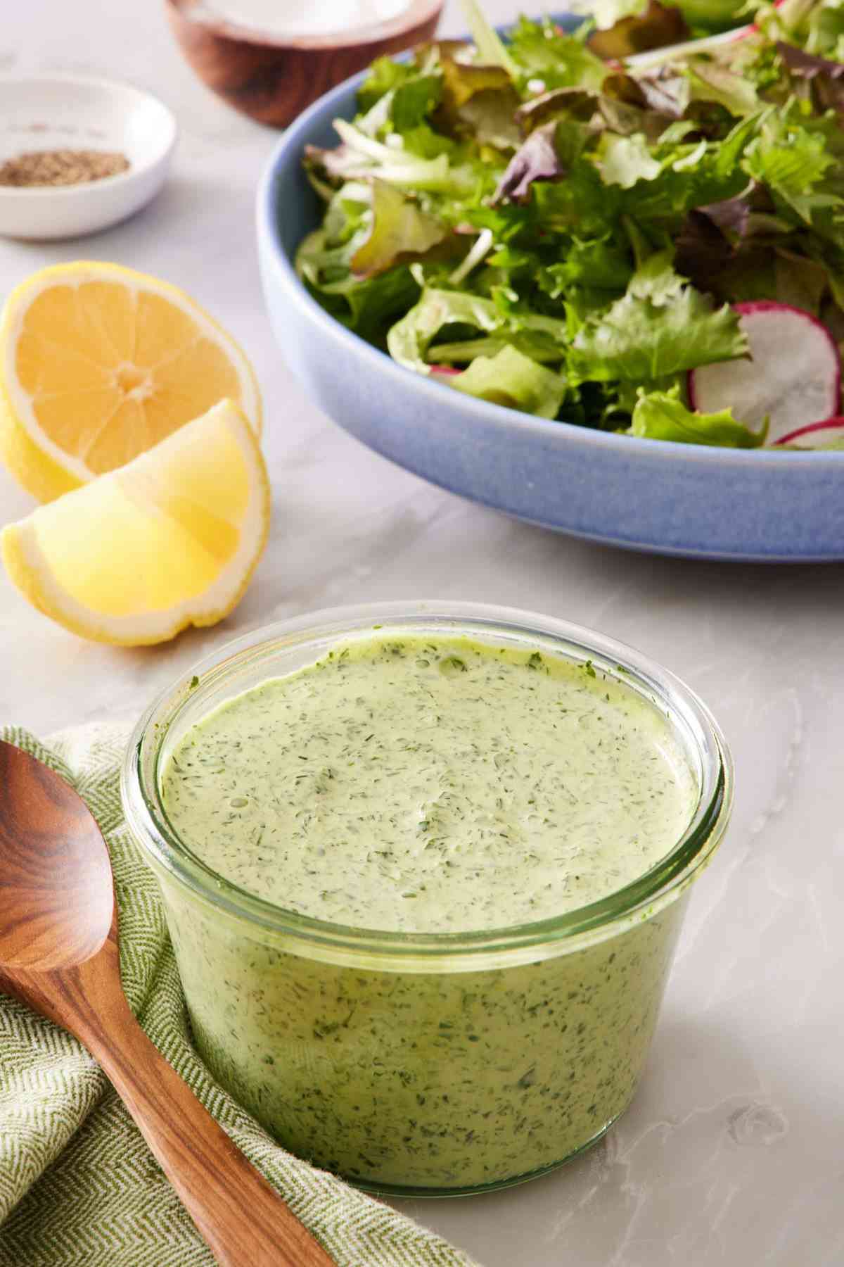 A glass jar filled with creamy green goddess dressing with sliced lemon and salad greens in the background.