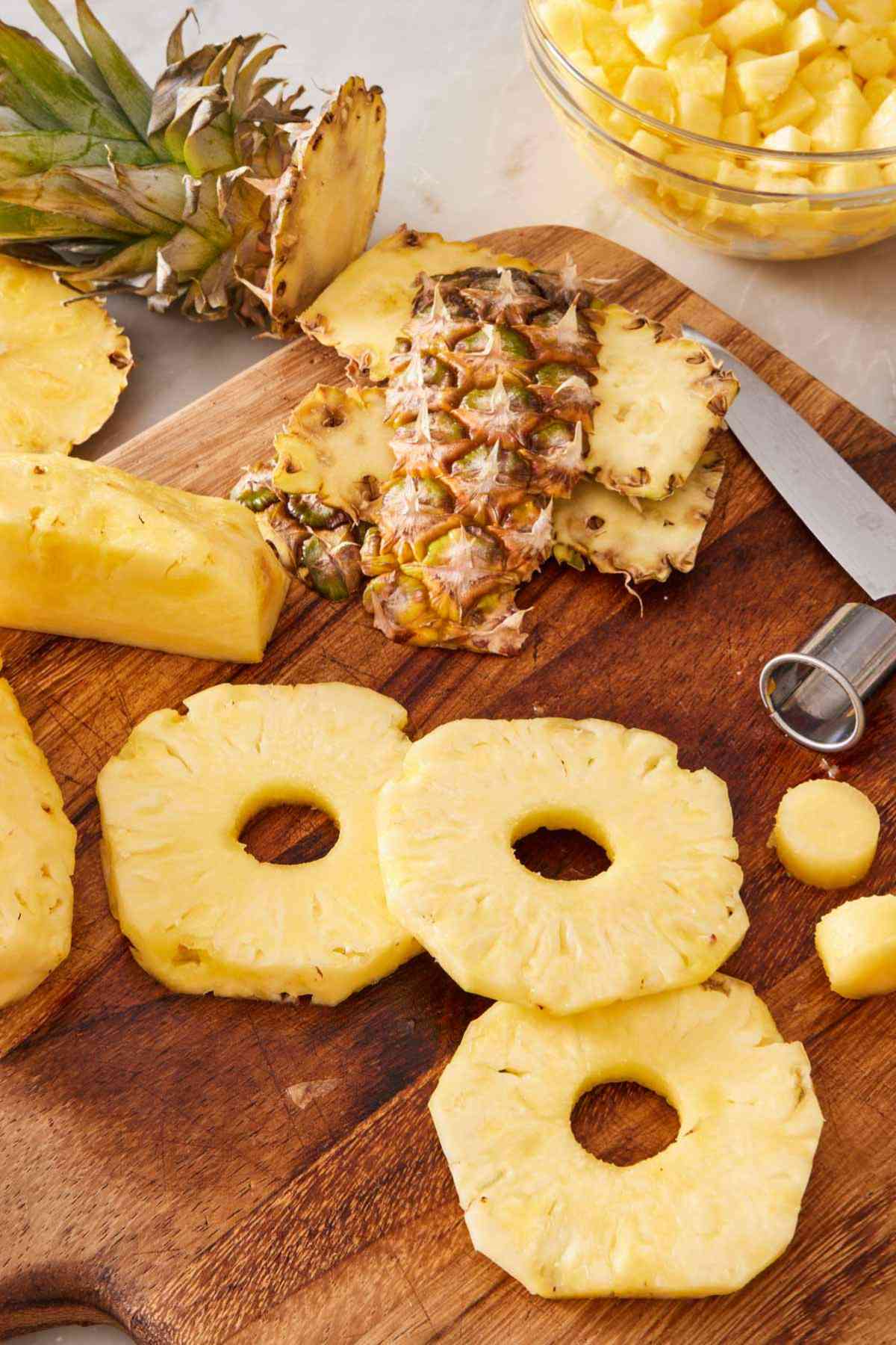Pineapple rings on a cutting board with the pineapple skin and diced pineapple in the background.