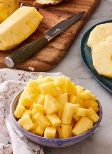 Diced pineapple in a bowl. Wooden cutting board with more pineapple in the background.