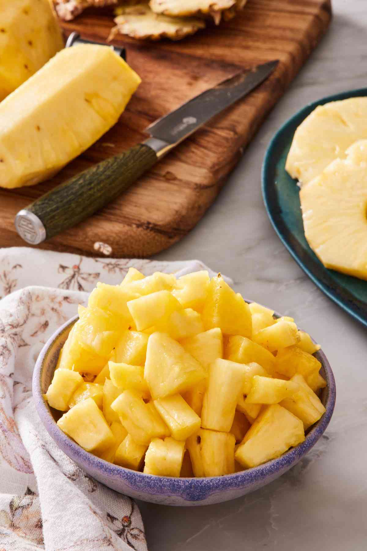 Diced pineapple in a bowl. Wooden cutting board with more pineapple in the background.