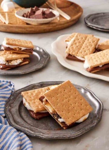 Two s'mores on a plate with more s'mores and ingredients for how to make s'mores in the background.