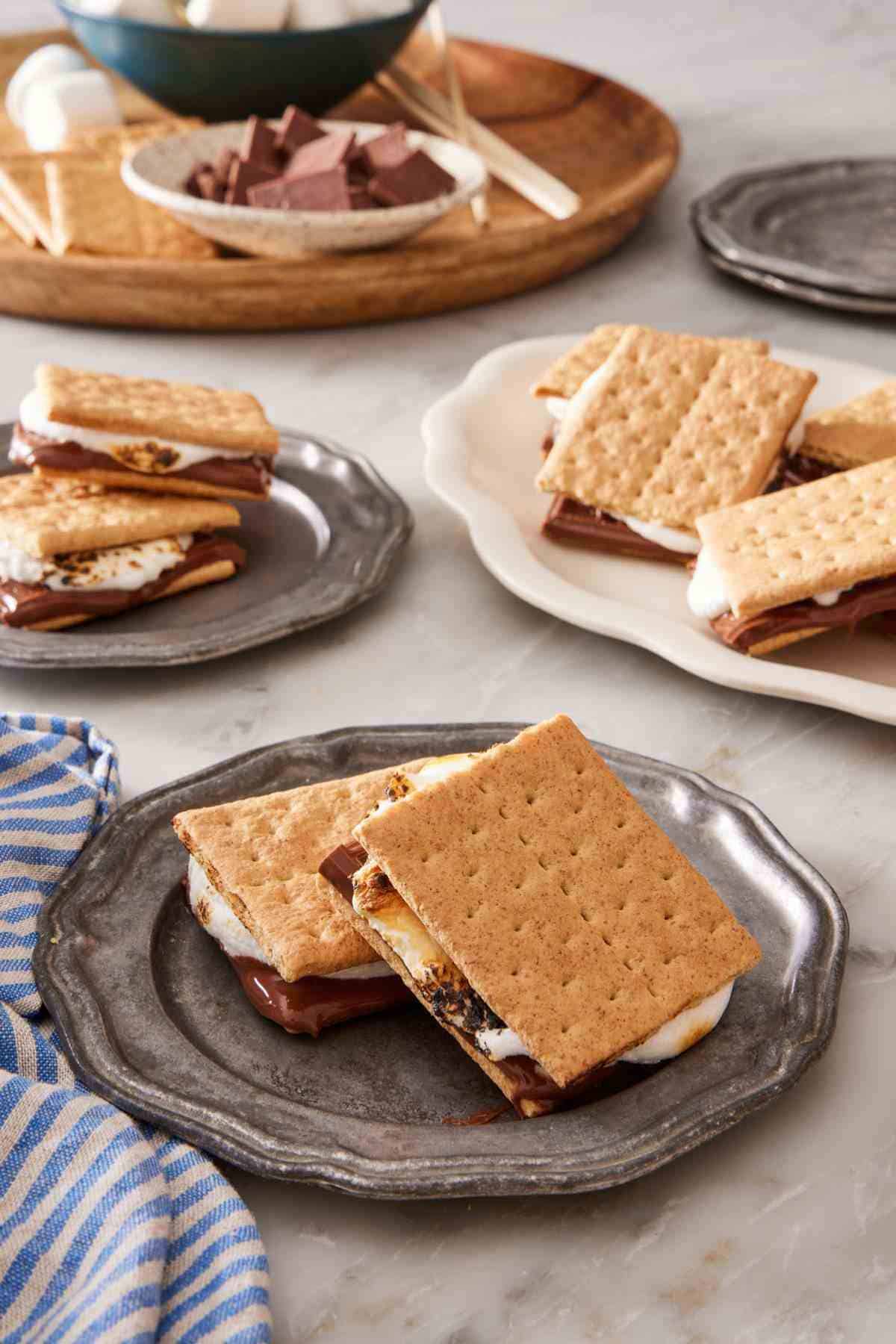 Two s'mores on a plate with more s'mores and ingredients for how to make s'mores in the background.