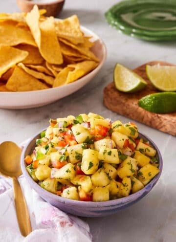 A heaped bowl of pineapple salsa on a countertop with lime wedges and chips in the background.
