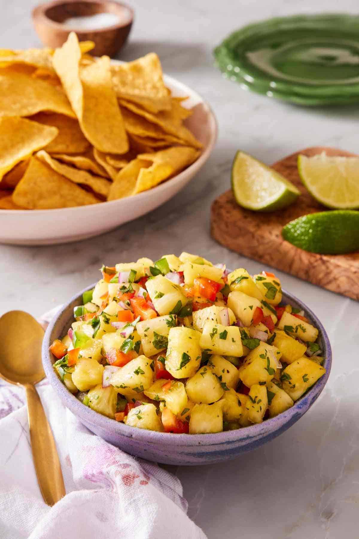 A heaped bowl of pineapple salsa on a countertop with lime wedges and chips in the background.