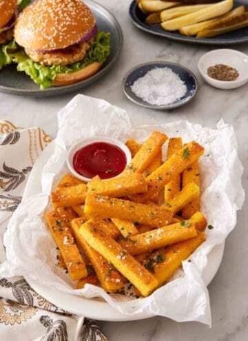 A dish of polenta fries on a paper towel with a burger in the background.