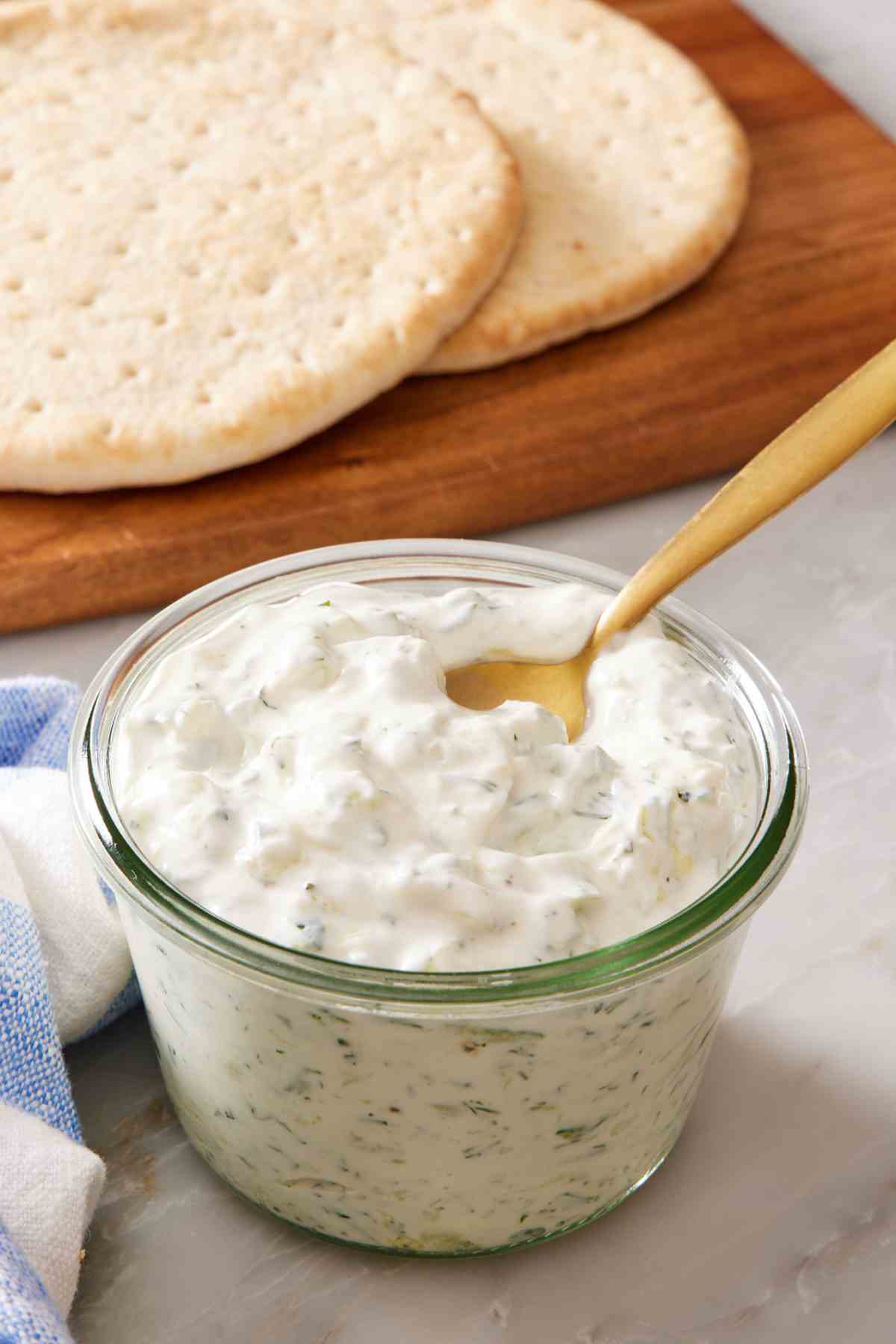 A small glass jar with tzatziki, with pita on a cutting board in the background.