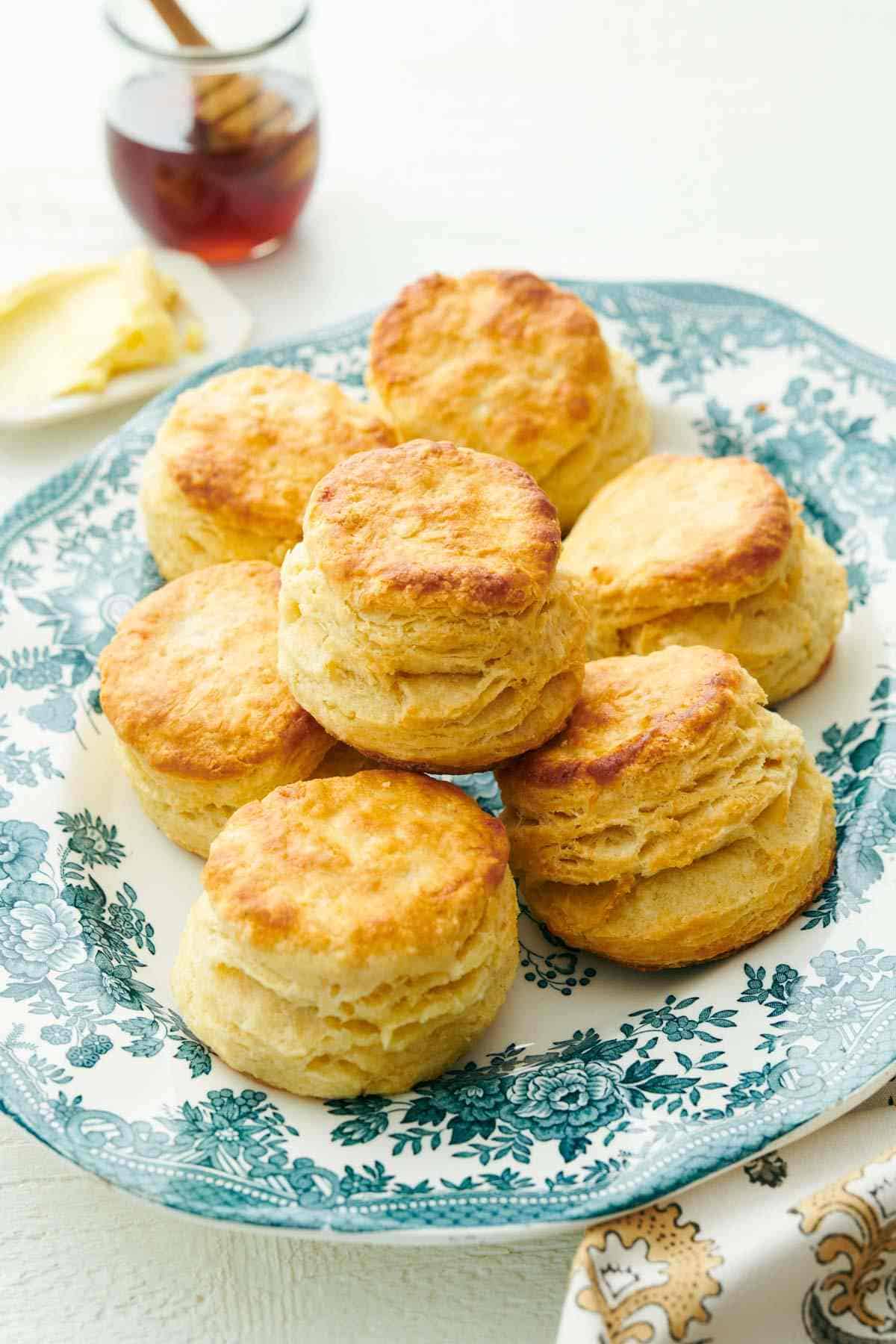 A patterned platter with several buttermilk biscuits with honey and butter in the background.