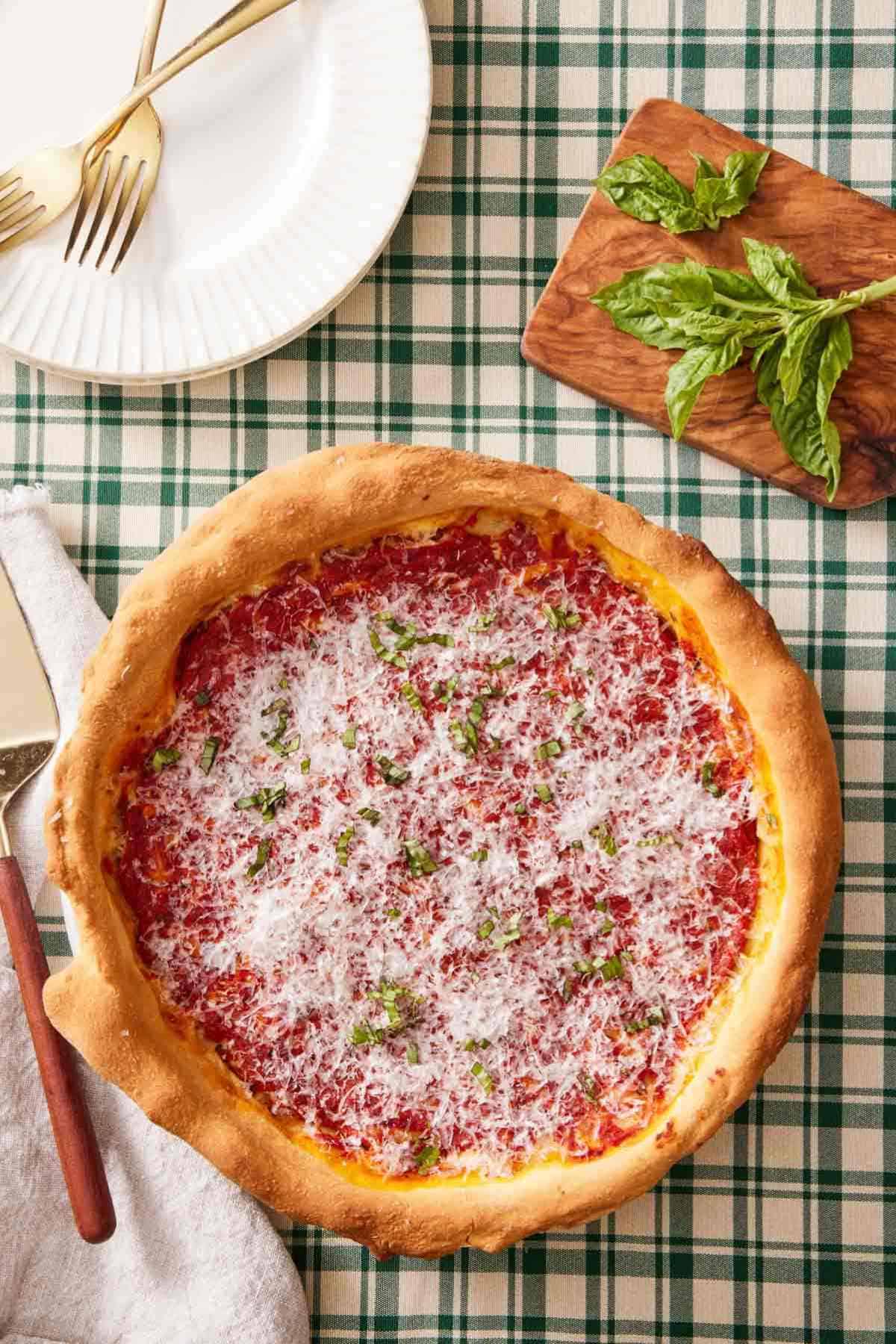 Overhead shot of a deep dish pizza on a checkered table linen with fresh basil and serving plates around it.