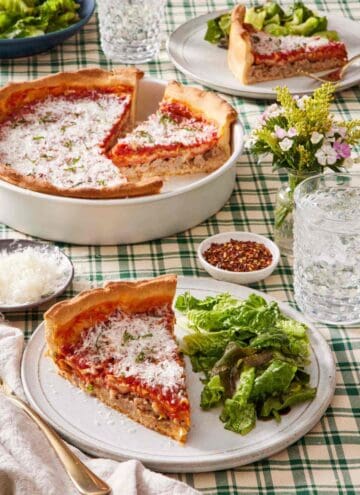 A slice of deep dish pizza on a plate with a side salad. A drink, dishes of red pepper flakes and shredded parmesan cheese, the rest of the pizza, and another slice of pizza, are also in frame.