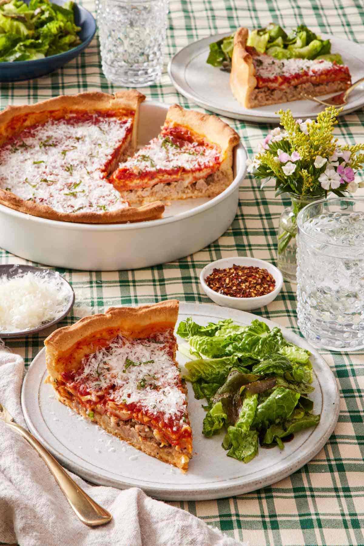 A slice of deep dish pizza on a plate with a side salad. A drink, dishes of red pepper flakes and shredded parmesan cheese, the rest of the pizza, and another slice of pizza, are also in frame.