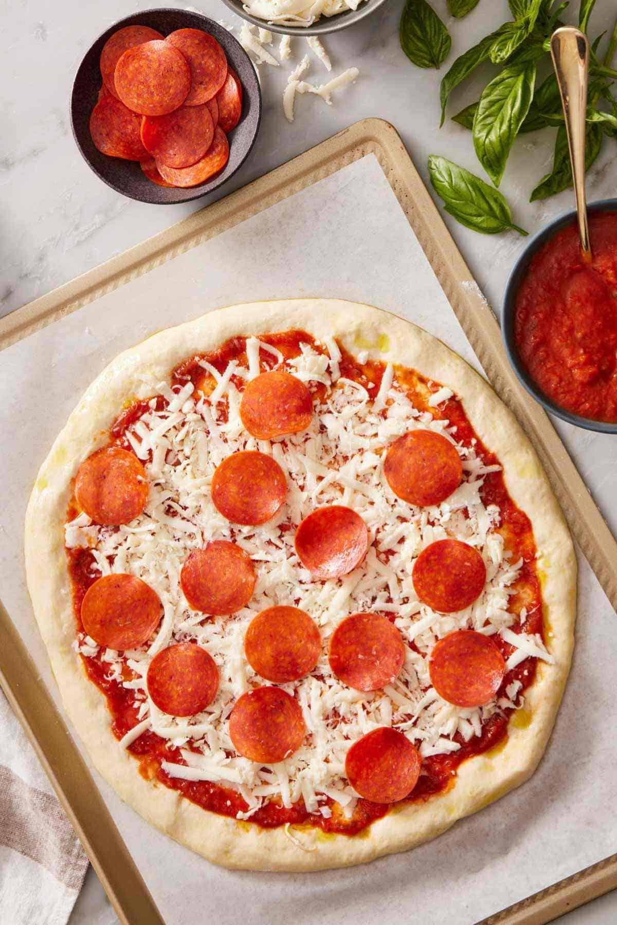 Overhead shot of a pepperoni pizza before baking on a baking sheet, with dishes of pizza sauce and pepperoni alongside.
