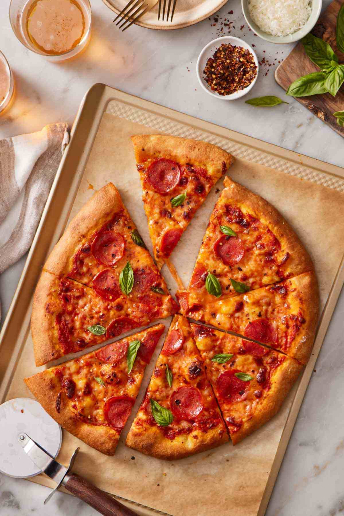 Overhead shot of a sliced homemade pepperoni pizza with chili flakes, shredded cheese, and drinks alongside.